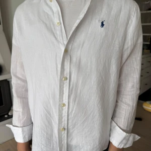 Vit linneskjorta från Polo Ralph Lauren - 14-16 år, L. Klassisk vit skjorta i linne från Polo Ralph Lauren med blå broderad logga på bröstet. Skjortan har långa ärmar, knappar framtill och en avslappnad passform. Perfekt för en stilren och fräsch look. 
