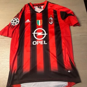 AC Milan fotbollströja Kaka 22 - Säljer en klassisk AC Milan fotbollströja med Kaka och nummer 22 på ryggen. Tröjan är röd och svart med vertikala ränder, Adidas-logga, klubbmärke och Opel som sponsor på framsidan. Kortärmad modell med Champions League-märke på ärmen.