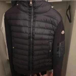 Navy blue cardigan från Moncler - Säljer en mörkblå dunjacka från Moncler med stickade ärmar och quiltad front. Jackan har dragkedja framtill, hög krage och en Moncler-logga på ärmen. Perfekt för kyliga dagar och riktigt snygg till vardags. Snabba affärer funkar!