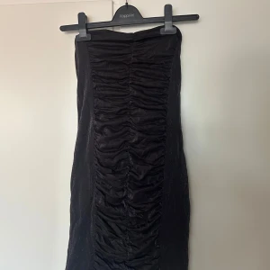 Svart axelbandslös omlottklänning - Säljer en svart axelbandslös klänning med snygg rynkad detalj framtill. Klänningen har en omlottliknande effekt och är figurnära med en glansig finish. Perfekt för dig som vill sticka ut med en elegant och stilren look.