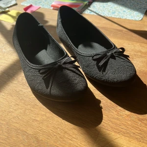 Svarta ballerinaskor med rosett från XIT - Svarta ballerinaskor från XIT med söt rosettdetalj framtill. Skorna har en rund tå och är täckta i ett diskret spetsliknande mönster. Perfekta för dig som gillar en klassisk och stilren look.