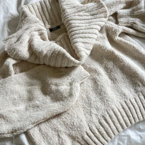 Beige stickad kofta med offshoulder krage - Mysig beige stickad kofta med ribbad offshoulder krage. Perfekt för lager-på-lager och kyliga dagar. Tjockt garn och långa ärmar ger en avslappnad och varm känsla.