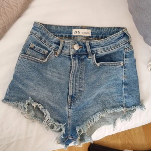 Blå jeansshorts med fransar från Zara - Säljer ett par klassiska blå jeansshorts från Zara med fransiga benslut och femficksmodell. Shortsen har hög midja och råa kanter för en trendig look. Perfekta till sommaren och lätta att matcha med olika toppar.