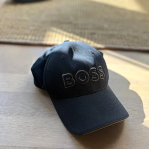 BOSS keps - Snygg blå keps från BOSS med broderad logotyp framtill i guld. Justerbart metallspänne baktill för perfekt passform. Klassisk böjd skärm och stilren design, passar till många tillfällen.