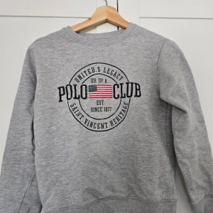 Grå sweatshirt från Polo Club Saint Vincent - Säljer en klassisk grå sweatshirt från Polo Club Saint Vincent med rund halsringning och tryckt logga framtill. Tröjan har långa ärmar och är tillverkad i mjukt material.