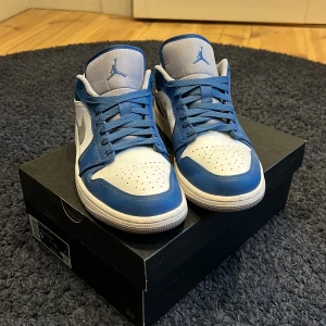 Nike Air Jordan 1 Low True Blue - Säljer ett par Nike Air Jordan 1 Low i färgen True Blue/Cement Grey-White. Skorna har blå och vita paneler med grå detaljer, klassisk snörning och rund tå. Perfekta sneakers för dig som gillar streetwear och ikonisk design.