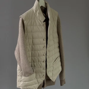  Suitsupply dunväst - Stilren beige dunväst med quiltat mönster och knappar framtill. Västen har ståkrage och två fickor på sidorna. Perfekt att slänga över en tröja för extra värme och snygg lager-på-lager-look.