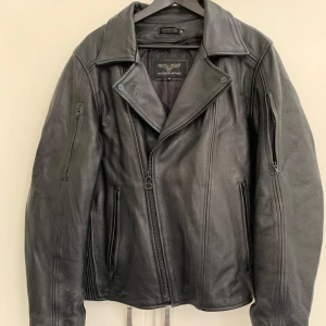 Svart skinnjacka med dragkedjor - Säljer en klassisk svart skinnjacka från Wilson Leather med asymmetrisk dragkedja och flera fickor med dragkedjor. Jackan har bred krage och långa ärmar, perfekt för en cool och tidlös stil.