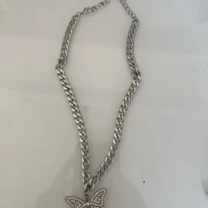 Silverfärgat halsband med fjärilshänge - Chunky silverfärgat halsband med ett glittrigt fjärilshänge dekorerat med små stenar. Kedjan är grov och ger ett trendigt intryck, perfekt för att lyfta din outfit.
