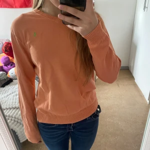 Persikofärgad tröja från Ralph Lauren - Säljer en persikofärgad långärmad tröja från Ralph Lauren med rund halsringning och diskret grön logga på bröstet. Tröjan har ribbade muddar vid ärmslut och nederkant. Perfekt för en avslappnad stil.