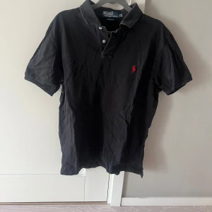 Svart pikétröja från Polo Ralph Lauren - Klassisk svart pikétröja från Polo Ralph Lauren med röd broderad logga på bröstet. Slim fit-modell med korta ärmar och två knappar vid kragen. Perfekt för en stilren och tidlös look.
