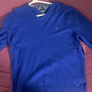 Blå v-ringad tröja från Tommy Hilfiger - Säljer en blå v-ringad tröja från Tommy Hilfiger i pima cotton cashmere. Tröjan har lång ärm och en liten broderad logga på bröstet. Perfekt för dig som gillar klassisk stil och mjuka material. Storlek lx men passar även L.