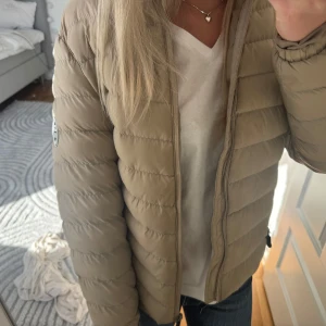 Beige pufferjacka med huva från Svea - Säljer en beige pufferjacka med dragkedja. Jackan har quiltad design och har vinsten polo märke på ärmen. Perfekt för kyligare dagar och har en klassisk, enkel look.