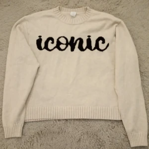 Beige stickad tröja med svart text från Lindex - Säljer en beige stickad tröja från Lindex med svart text 'iconic' framtill. Tröjan har rund halsringning, långa ärmar och ribbade muddar vid ärmslut och nederkant. Perfekt för en avslappnad och trendig look.