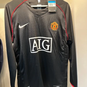 Manchester United långärmad fotbollströja - Säljer en svart långärmad Manchester United fotbollströja från Nike med AIG som sponsor på bröstet. Tröjan har röda detaljer längs axlarna och klubbmärket broderat på vänster bröst. Materialet är lätt och andas, perfekt för match eller träning. Alla storlekar finns 