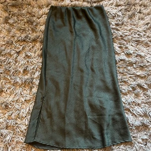 Mörkgrön satinkjol med slits - Säljer en lång, mörkgrön kjol i satin med diskret struktur och två små slits på var sin sida. Kjolen har en elegant, rak passform och faller snyggt längs kroppen. Perfekt för dig som gillar stilrena plagg med en twist.
