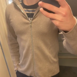Beige ribbad ziptröja från LUNG - Säljer en beige ribbad ziptröja från LUNG med hög krage och hel dragkedja framtill. Tröjan har lång ärm och är i ett mjukt, stretchigt material som sitter snyggt på kroppen. Perfekt för lager-på-lager och enkel att matcha. Använd några gånger, ny skick. Passar som en mindre large