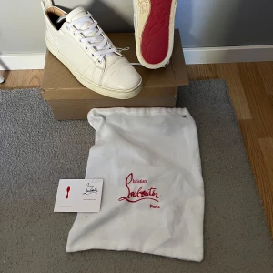 Vita sneakers från Christian Louboutin - Säljer ett par vita sneakers från Christian Louboutin med klassisk röd sula. Skorna har rund tå, snörning och är tillverkade i skinn. Perfekta för dig som gillar stilrena och exklusiva sneakers. Tar emot byten eller skicka prisförslag