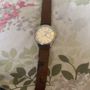 Regal herrklocka med brun läderrem - Stilren klocka från Regal med vit urtavla, silverfärgat boett och bruna skinnband. Urtavlan har tydliga markeringar och en extra liten sekundvisare. Modellnummer R1685. Perfekt accessoar för dig som gillar klassisk design. Du ansvara för leverans 