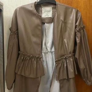 Brun kappa i skinnimitation från Samdan - Säljer en brun kappa i skinnimitation från Samdan. Jackan har långa ärmar med rynkade detaljer vid armbågar och midja samt knytband framtill. Perfekt för dig som vill ha en trendig och stilren look.