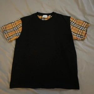 Unik t-shirt med Burberry-stil ärmar – trendig streetwear! - Säljer en riktigt snygg och unik svart t-shirt med klassiska rutiga Burberry-inspirerade ärmar. Väldigt trendig design som sticker ut utan att vara för mycket – perfekt för dig som gillar streetwear med stil!                                                                                       ✅ I mycket fint skick ✅ Storlek: M men passar även S ✅ Mjuk och bekväm passform ✅ Perfekt till våren/sommaren eller under en hoodie