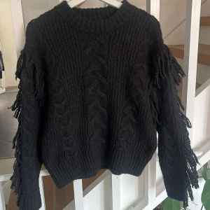 Svart stickad tröja med fransar från Gina Tricot - Säljer en svart stickad tröja från Gina Tricot i storlek XS. Tröjan har flätstickat mönster och långa ärmar med fransdetaljer längs sidorna. Perfekt för dig som vill ha en unik och trendig look.