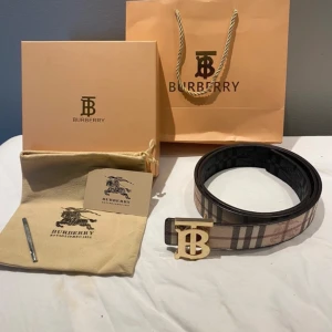 Burberry bälte med rutigt mönster och guldfärgat spänne - Säljer ett Burberry bälte med klassiskt rutigt mönster i beige, svart, vitt och rött. Bältet har ett stort guldfärgat spänne med Burberrys logotyp. Perfekt accessoar för att lyfta din outfit.