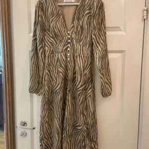 Zebramönstrad långklänning från Adoore - Snygg långklänning från Adoore  med zebramönster i beige och svart. Klänningen har v-ringning, knappar framtill och långa ärmar. Perfekt för dig som vill sticka ut med ett djurmönster och en elegant siluett., den har en liten tråd om har gått upp men ingeting så syns!🩷