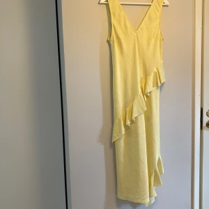 Gul klänning med volang - Säljer en ljusgul ärmlös omlottklänning med v-ringning och volangdetaljer längs framsidan. Klänningen har en elegant och somrig känsla, perfekt för varma dagar. Materialet är lätt och har en diskret struktur.