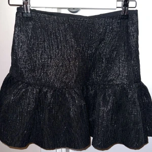 Svart glittrig kjol från Zara - Snygg svart glittrig kjol från Zara med volangdetalj. Kjolen har en dragkedja baktill för enkel på- och avtagning. Perfekt för att ge lite extra bling till din outfit!