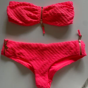Rosa bandeau bikini med dragkedjedetaljer - Säljer en snygg rosa bandeau bikini med struktur och dragkedjor både på toppen och trosorna. Bikinin har en modern look med texturerat tyg och guldiga dragkedjor som detalj. Perfekt för sommaren!