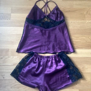 Lila satin pyjamasset med spetsdetaljer - Säljer ett lila pyjamasset i satin med svarta spetsdetaljer. Linnet har tunna axelband och transparent spets framtill, och shortsen har matchande spets längs sidorna. Perfekt för mysiga kvällar hemma.