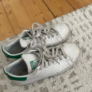 Adidas Stan Smith vita sneakers med gröna detaljer - Klassiska vita Adidas Stan Smith sneakers med gröna detaljer på hälen och tungan. Skorna har snörning, rund tå och platt sula. Tillverkade i skinn med diskreta perforeringar på sidorna för ventilation. Perfekta för en stilren och sportig look.
