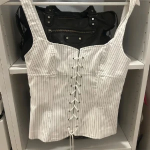 Vit randig korsettopp med snörning - Säljer en vit korsettopp med tunna svarta ränder och snörning framtill. Toppen har breda axelband och en figurnära passform som ger en snygg siluett. Perfekt att styla med jeans eller kjol för en trendig look.