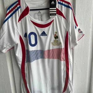 Frankrike fotbollströja Zidane #10 - Säljer en vit fransk landslagströja från Adidas med blå och röda detaljer. Nummer 10 och Zidane på ryggen, samt broderad landslagslogga och Adidas-märke på bröstet. Klassisk design med korta ärmar och rund hals.