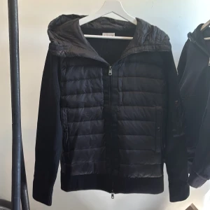 Moncler cardigan - Säljer en svart Moncler-jacka med quiltad front och huva. Jackan har dragkedja, ribbade muddar och är tillverkad i en mix av material med stickade ärmar och baksida. Klassisk Moncler-logga på ärmen.