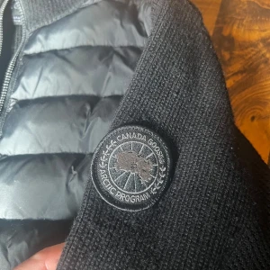 Svart hybridjacka från Canada Goose - Säljer en svart Canada Goose-jacka med quiltade partier framtill och stickade ärmar och sidor. Jackan har dragkedja och en rund patch på ärmen. Perfekt för kyliga dagar när du vill ha både stil och värme.