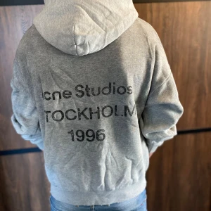 Grå hoodie från Acne Studios - Säljer en grå hoodie från Acne Studios med tryck på ryggen: 'Acne Studios STOCKHOLM 1996'. Klassisk modell med huva och ribbade muddar. Perfekt för en avslappnad stil. Hör av dig vid minsta fråga!