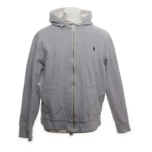Grå hoodie från Polo Ralph Lauren - Klassisk grå hoodie från Polo Ralph Lauren med dragkedja framtill och huva. Tröjan har långa ärmar, två fickor och en liten broderad logga på bröstet. Perfekt för en avslappnad stil.