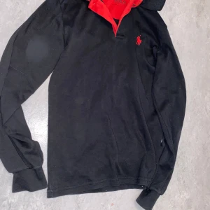  Polo Ralph Lauren Svart långärmad pikétröja  - Säljer en svart långärmad pikétröja från Polo Ralph Lauren med röd krage och röd broderad logga på bröstet. Klassisk modell med mjukt bomullsmaterial och snygga detaljer. Perfekt för dig som gillar stilrena plagg med en sportig touch.