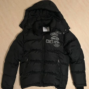 Svart pufferjacka från Mercier med huva - Svart pufferjacka från Mercier med huva och dragkedja. Jackan har flera broderade patches på bröstet och två fickor med dragkedja framtill. Perfekt för kyligare dagar.