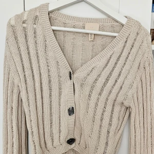 Beige croppad kofta från H&M - Säljer en beige croppad kofta från H&M med ribbat stickat material och stora knappar framtill. Tröjan har lång ärm och en avslappnad passform, perfekt att slänga över en topp.