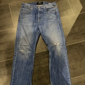 Blå jeansbyxor från Replay - Säljer ett par klassiska blå jeans från Replay med raka ben och diskreta slitningar framtill. Byxorna har fem fickor, knappgylf och detaljerad Replay-logga vid fickan. Perfekta för en avslappnad stil.