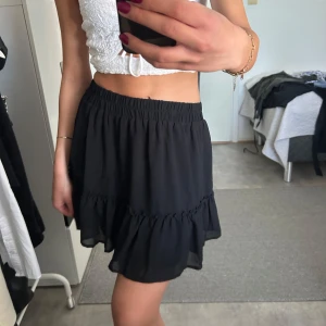 Svart volangkjol - Säljer en svart kortkjol med volangdetalj och resår i midjan. Kjolen har en luftig och flowy passform som gör den perfekt för varma dagar. Enkel att matcha med olika toppar för en trendig look.