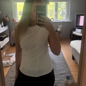 Vit omlottopp från Zara - Säljer en vit ärmlös omlottopp från Zara med snygg drapering över axeln. Toppen har en elegant och stilren look, perfekt att matcha med både jeans och kjol. Materialet känns mjukt och följsamt mot kroppen.