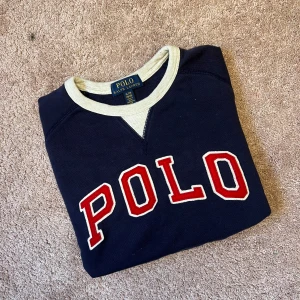 Marinblå sweatshirt från Polo Ralph Lauren - Säljer en marinblå sweatshirt från Polo Ralph Lauren med stora röda bokstäver 'POLO' framtill. Tröjan har vita och blå ribbade muddar vid ärmslut och nederkant samt kontrastfärgad krage.
