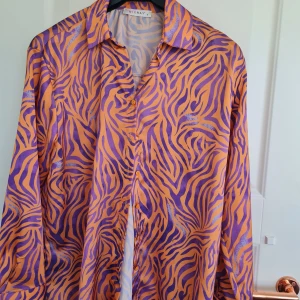 Orange och lila zebramönstrad skjorta från Mixray - Färgstark skjorta från Mixray i orange och lila med zebramönster. Skjortan har klassisk krage, långa ärmar och knäppning framtill. Perfekt för dig som vill sticka ut med en unik och trendig look.