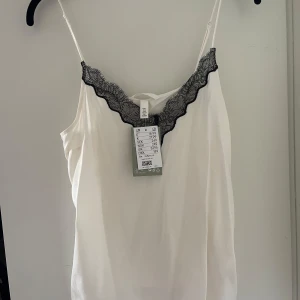 Vit satinlinne med svart spets  - Säljer en vit satintopp med svart spets från H&M med tunna axelband. Enkel och stilren design, perfekt till sommaren och fest