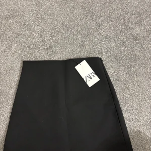 Svart minikjol från ZARA - Säljer en svart minikjol från ZARA. Kjolen har en rak passform och är längre på ena sidan. Helt oanvänd och med prislapp kvar. 