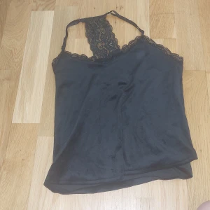 Svart linnetopp med spetsdetaljer från hunkemöller - Säljer en svart linnetopp med tunna axelband och fina spetsdetaljer både fram och bak. Toppen har en elegant v-ringning och är tillverkad i ett mjukt, glansigt material som känns lyxigt mot huden.